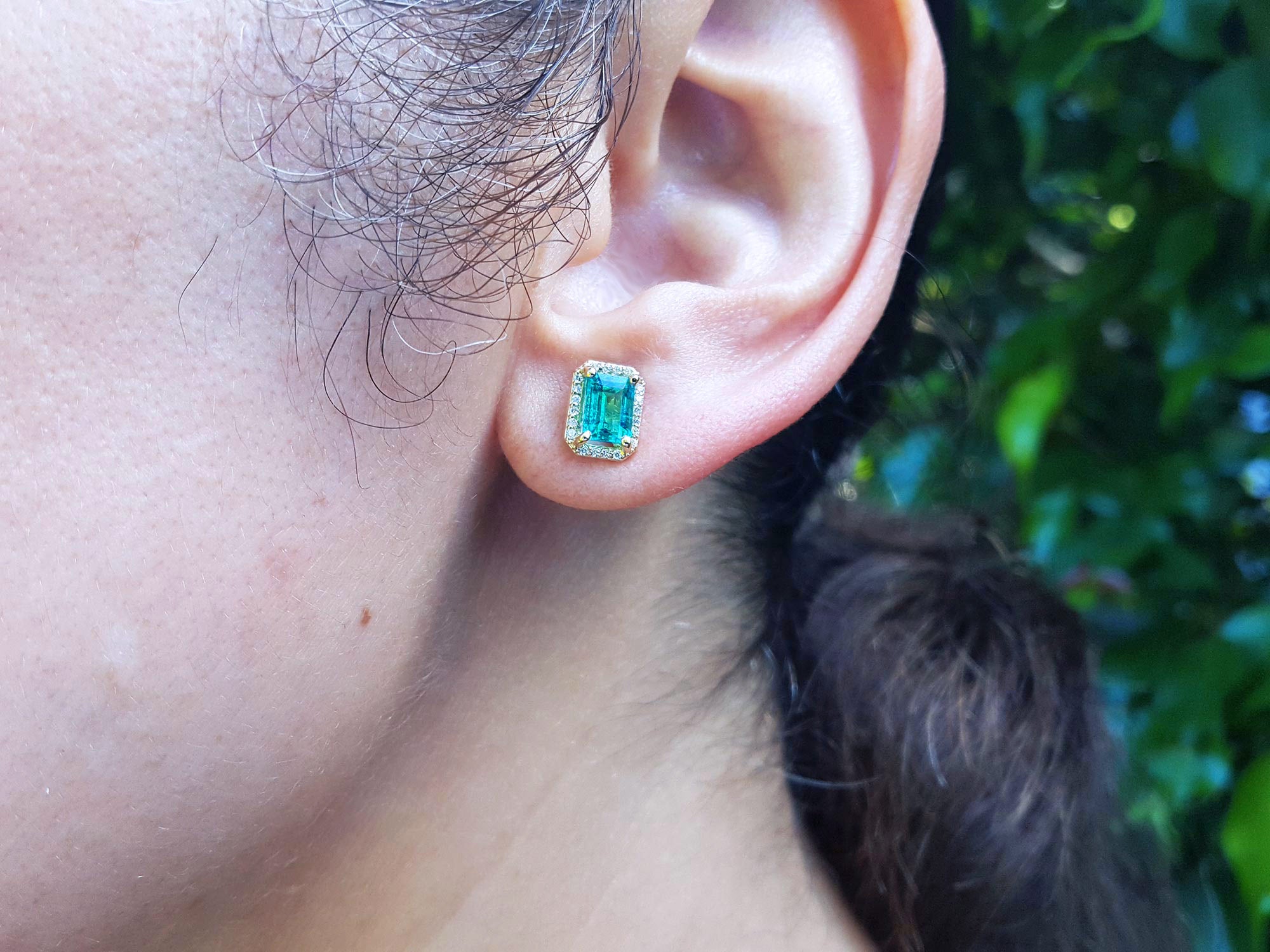 Close-up front view of an ear wearing a 14k gold emerald sud earring with a blurred green background