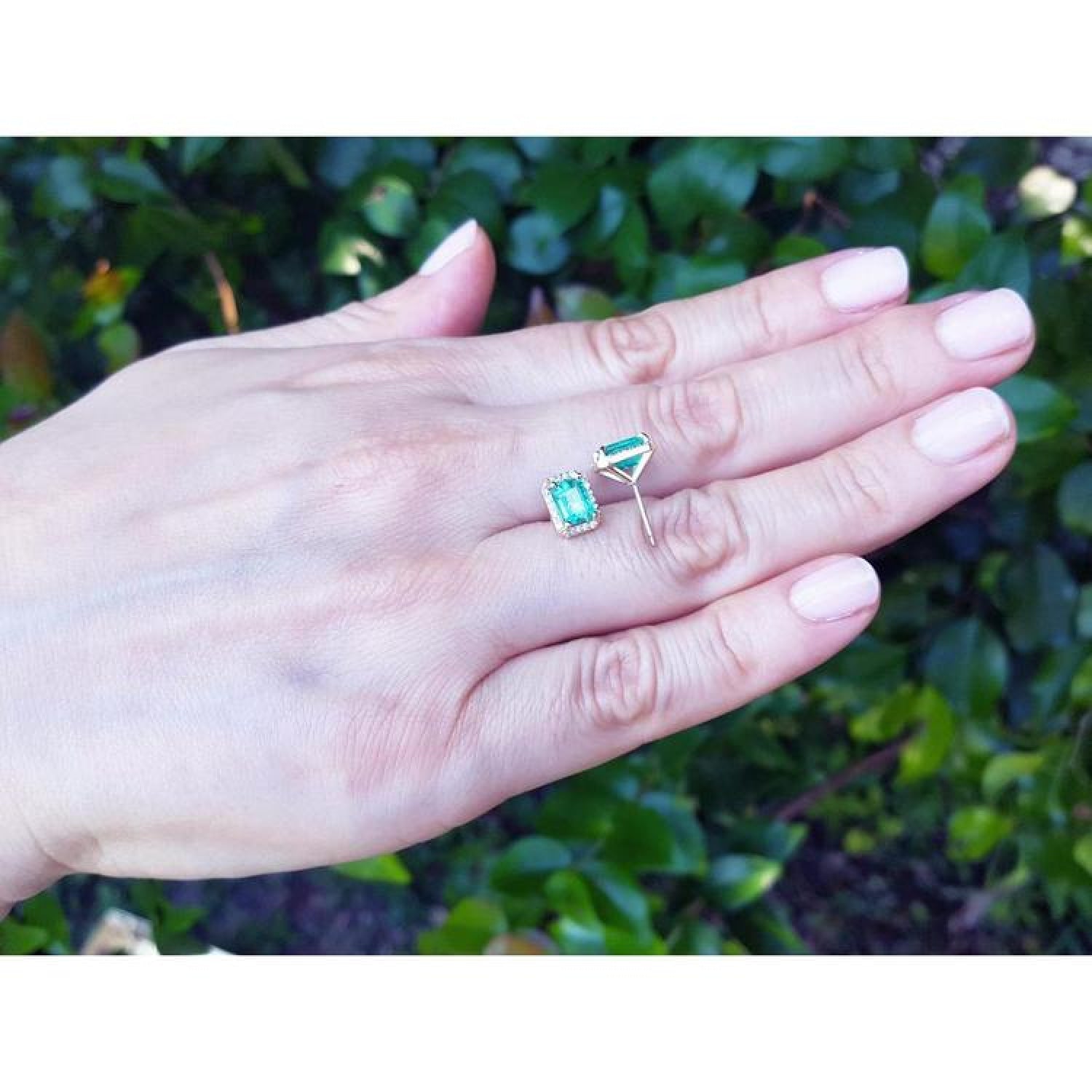 Emerald stud earrings with diamond halo in 14k yellow gold, displayed on a hand.