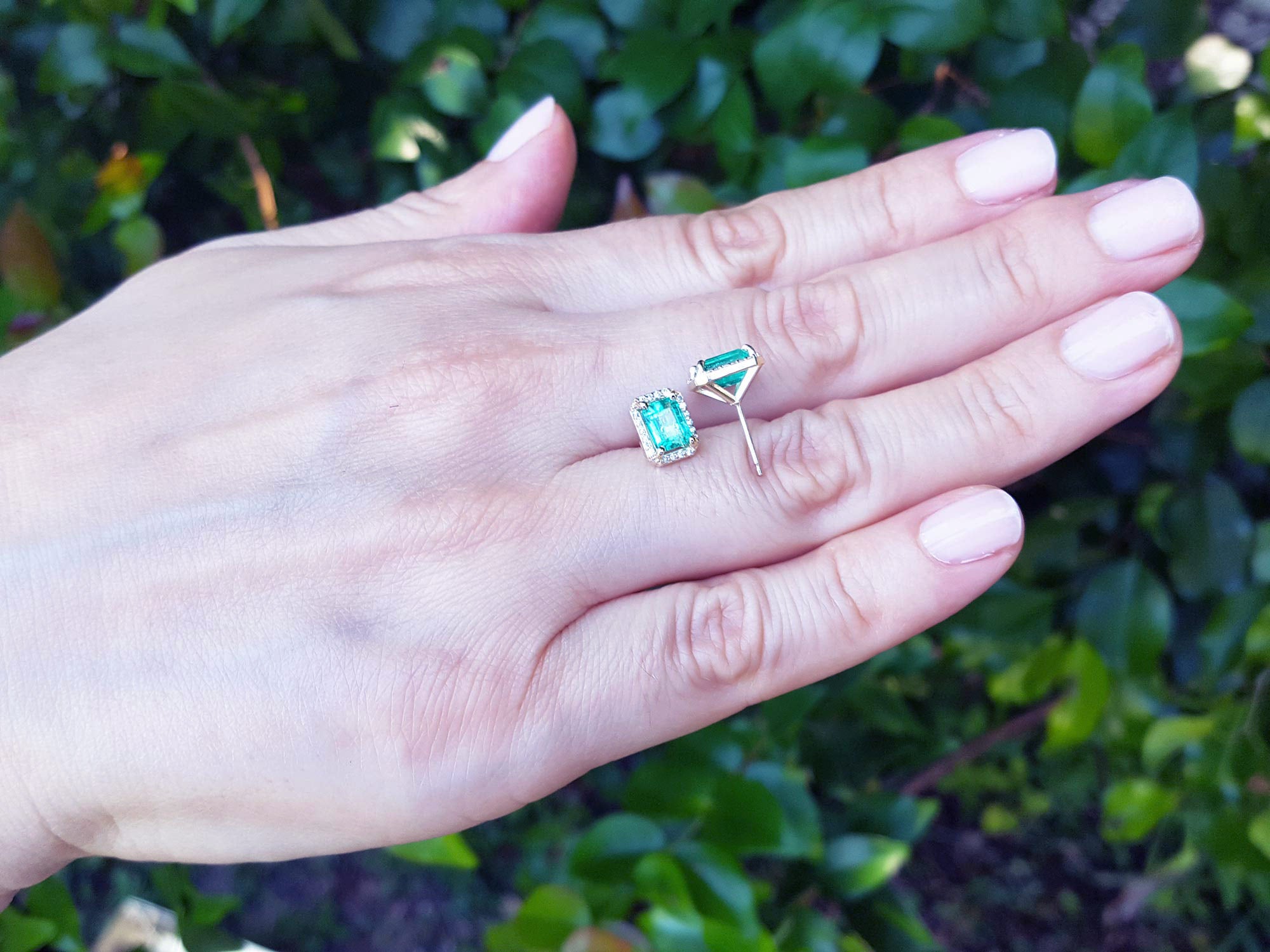 Yellow gold earrings with natural emeralds held in a hand against a blurred green background