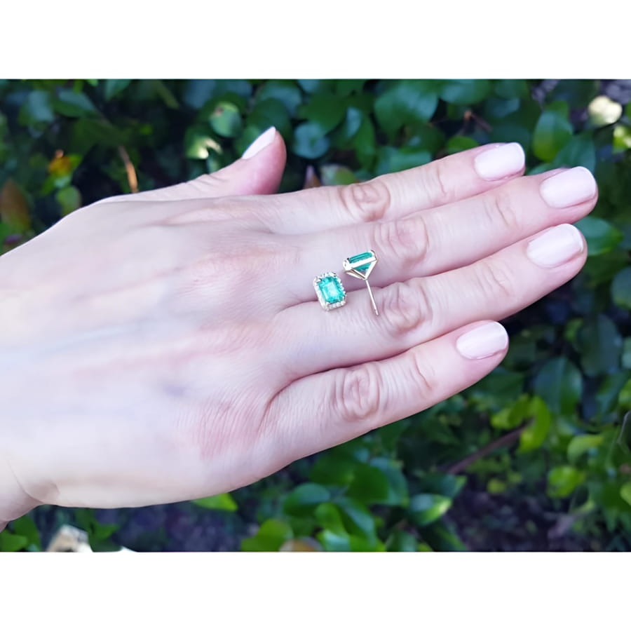 Emerald stud earrings with diamond halo in 14k yellow gold, displayed on a hand.