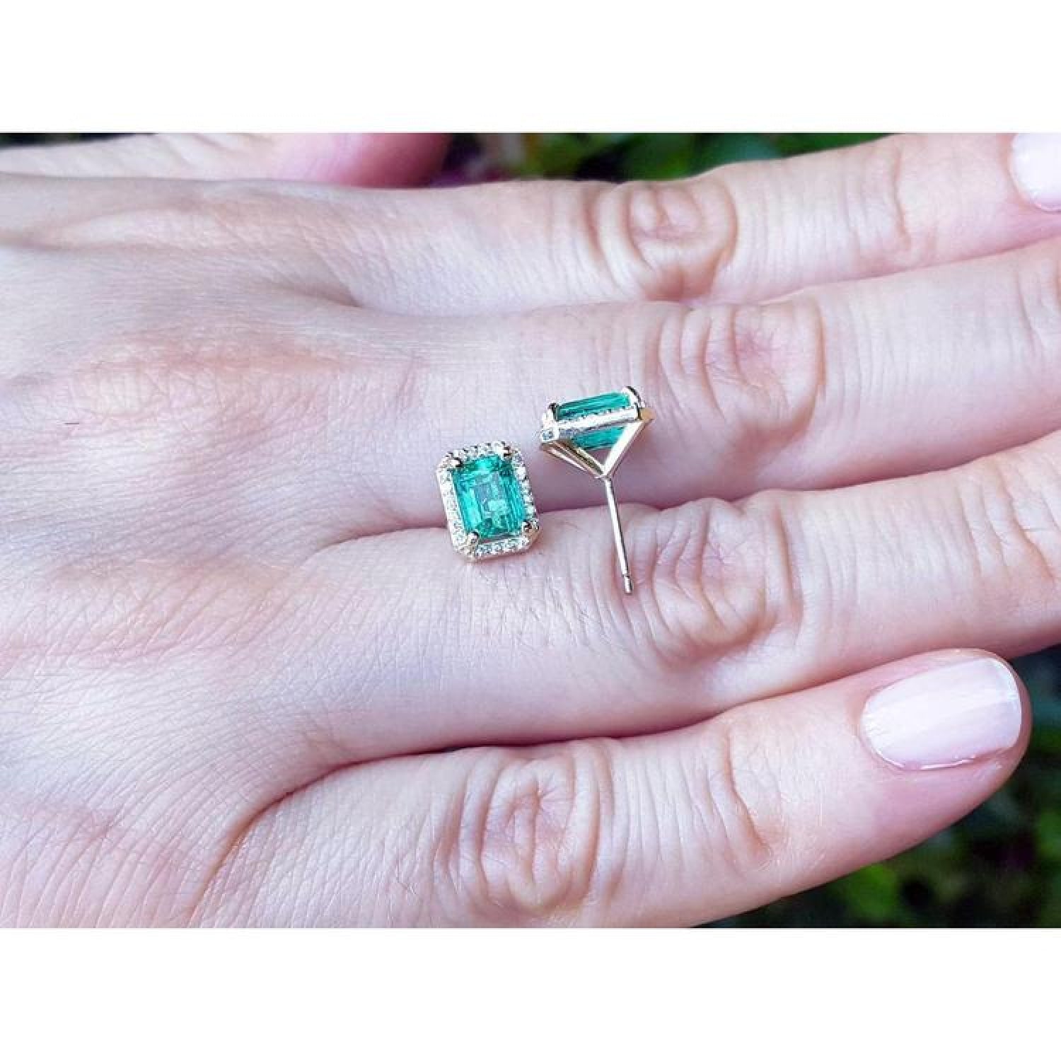Emerald stud earrings with diamond halo in 14k yellow gold, displayed on a hand.