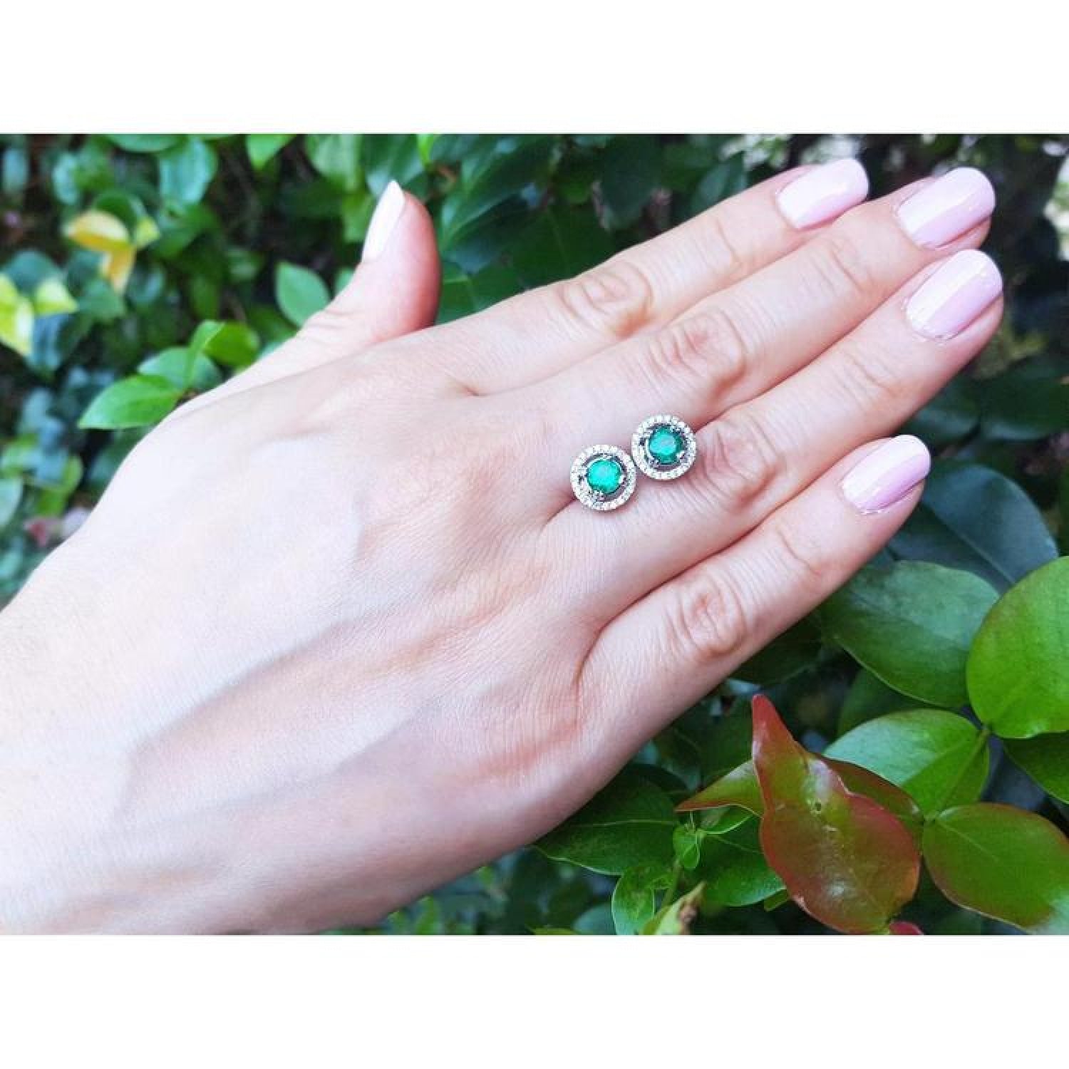 Halo emerald stud earrings with deep bluish-green 1.06 ct. emeralds encircled by diamonds on a hand.
