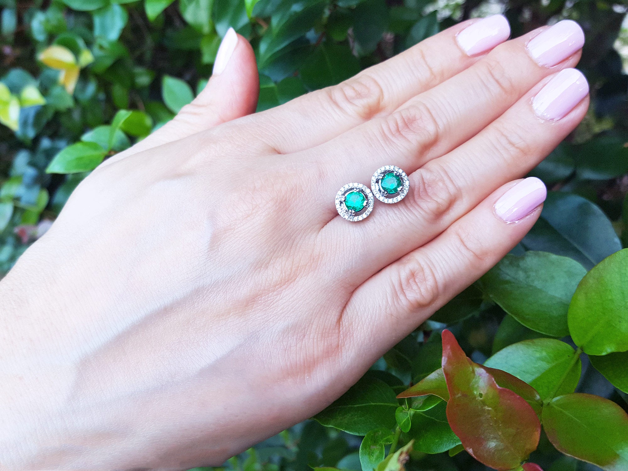 Hand displaying a pair of emerald stud earrings with a green leafy background