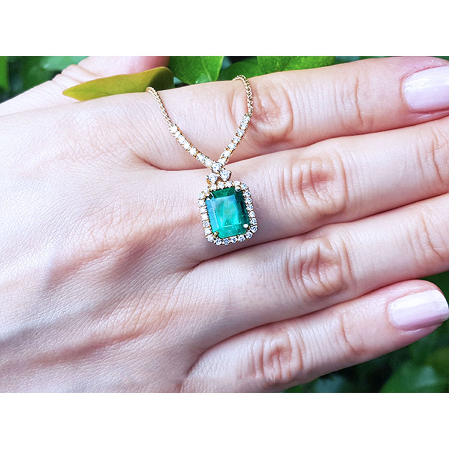 Emerald halo necklace with natural diamonds on a 14K yellow gold chain, held by a hand.