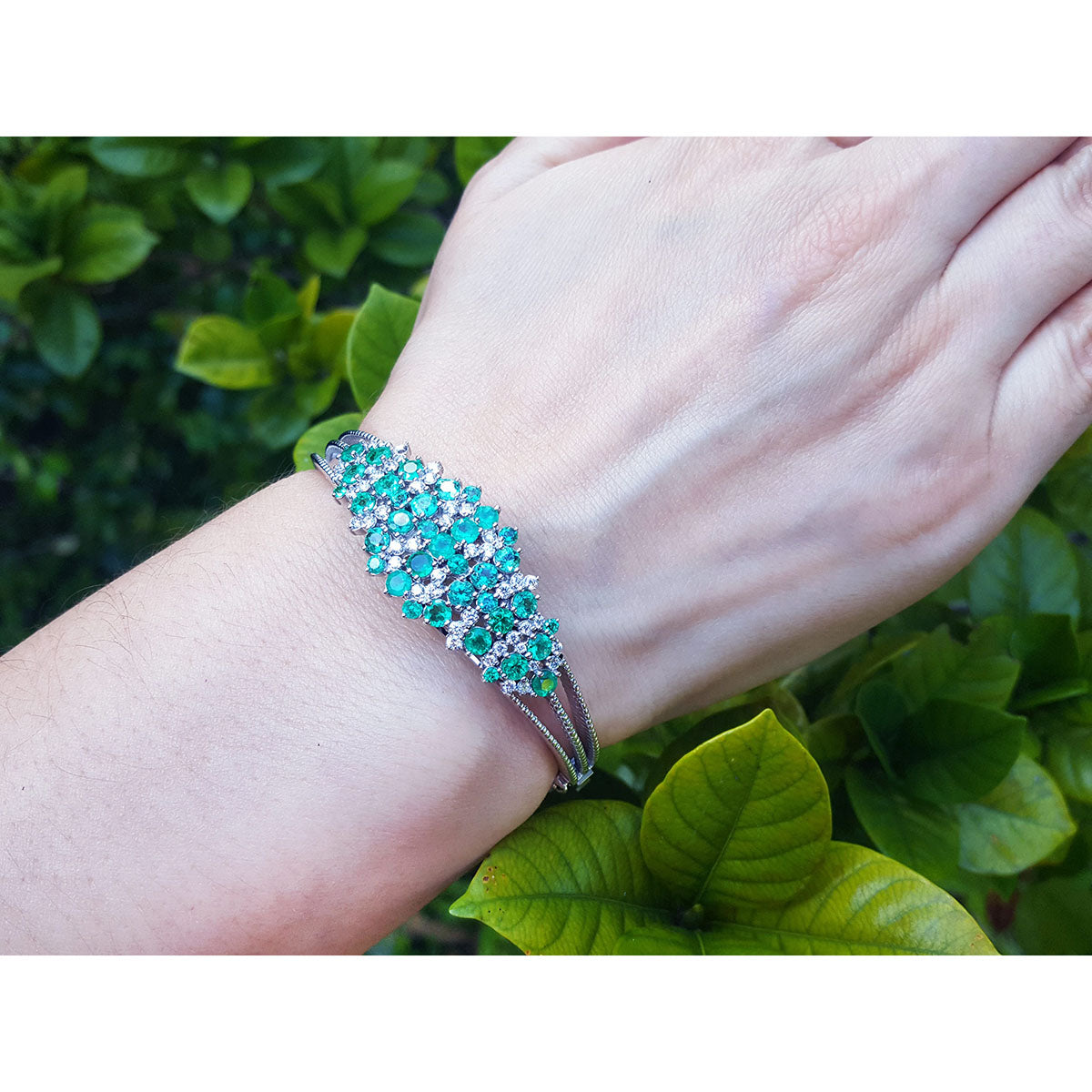 Emerald and diamonds bangle bracelet on a wrist with a green leafy background.