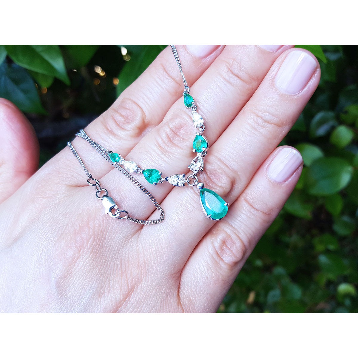 Hand holding White gold 14k Emerald necklaces pear shaped wheat style chain against a blurred green background