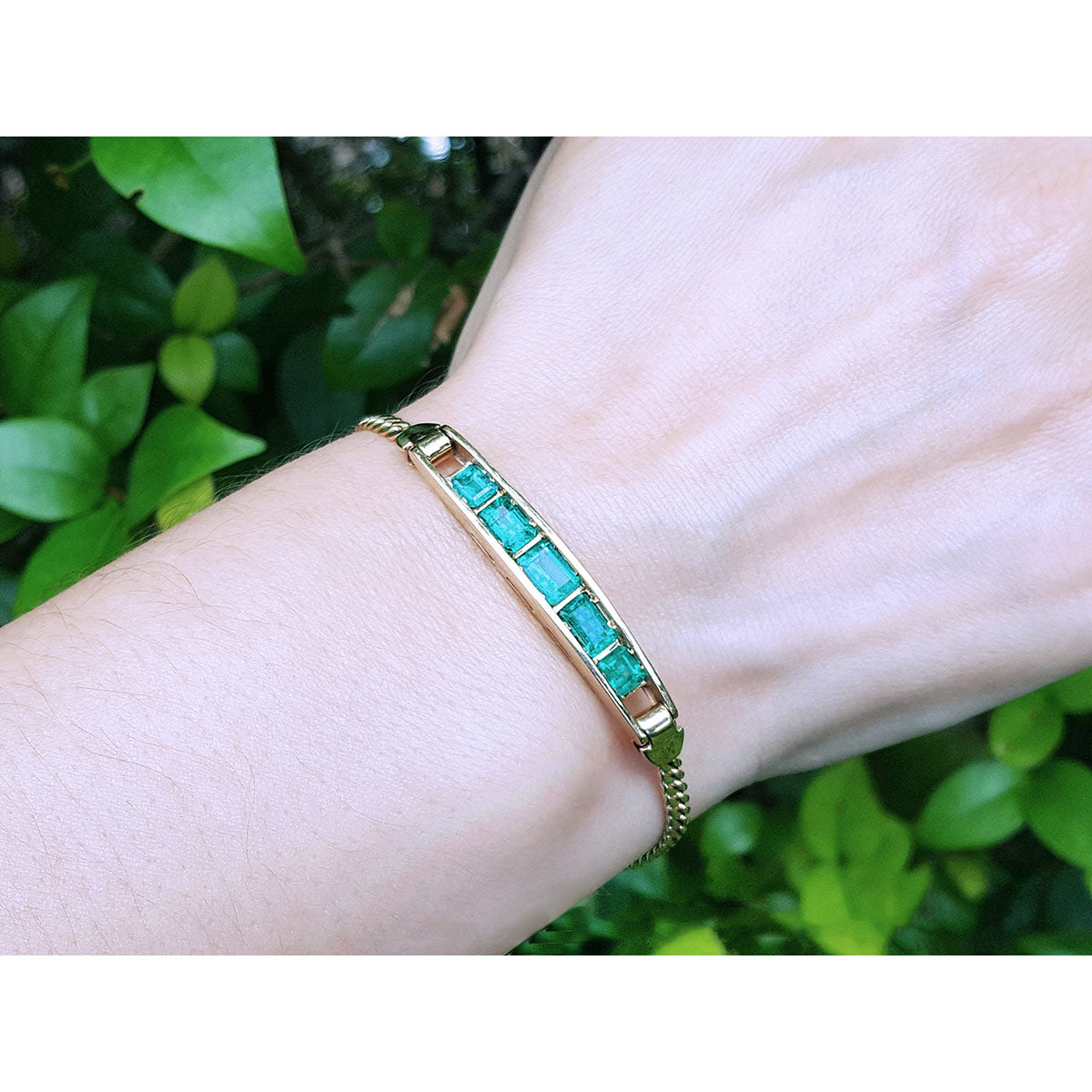 Gold bracelet with five emerald stones on a wrist against a green leafy background.