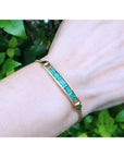 Gold bracelet with five emerald stones on a wrist against a green leafy background.