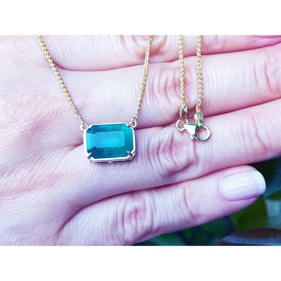 Emerald-cut Colombian emerald solitaire necklace in 18K yellow gold, held in a hand.