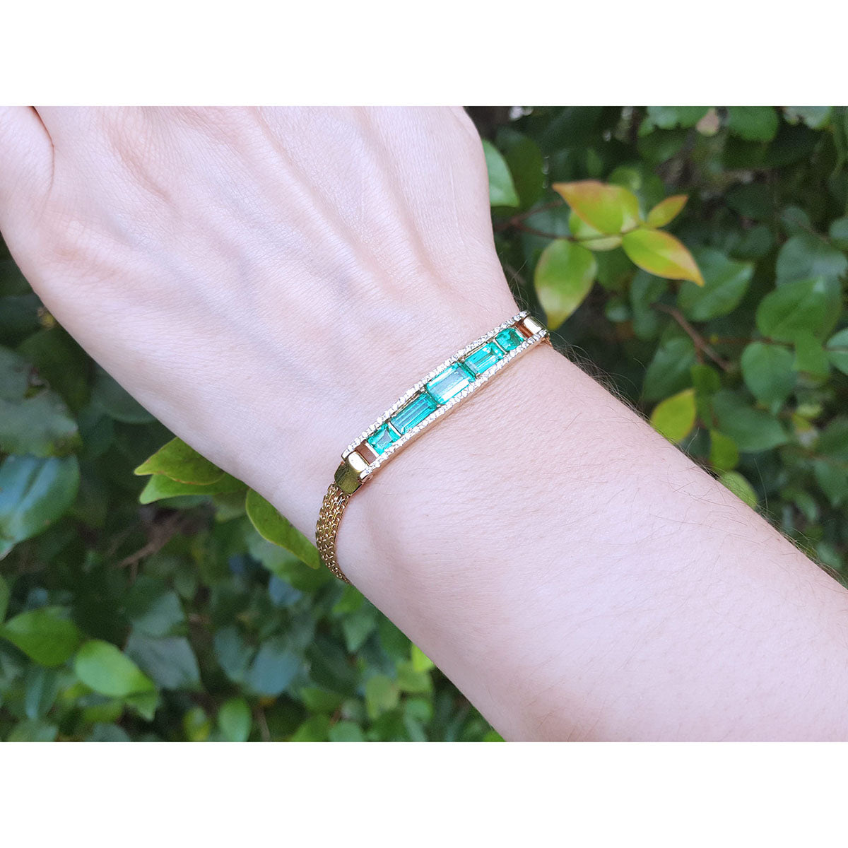 Gold bracelet with real colombian emeralds on a wrist against a green leafy background.