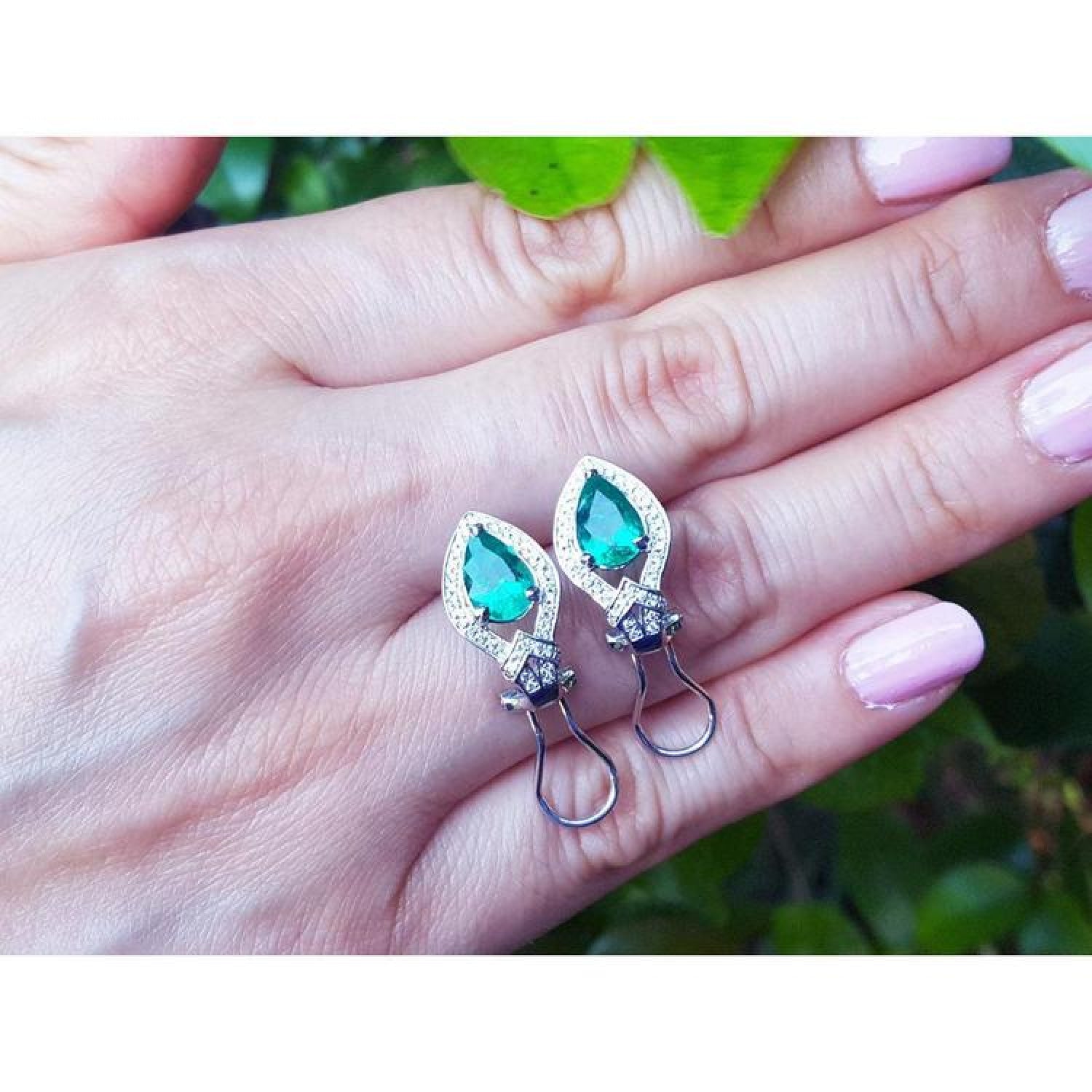 14K white gold clip/post earrings with pear-shaped Colombian emeralds and diamond accents on a hand.