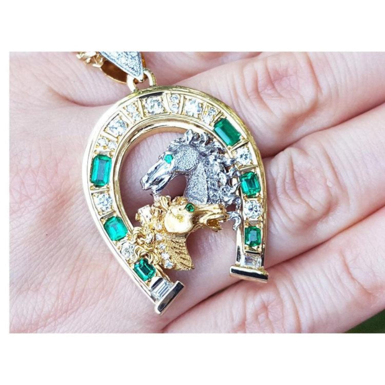18K yellow gold horseshoe pendant with emeralds, diamonds, and two horse heads exhibited on a hand.