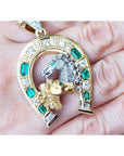 18K yellow gold horseshoe pendant with emeralds, diamonds, and two horse heads exhibited on a hand.