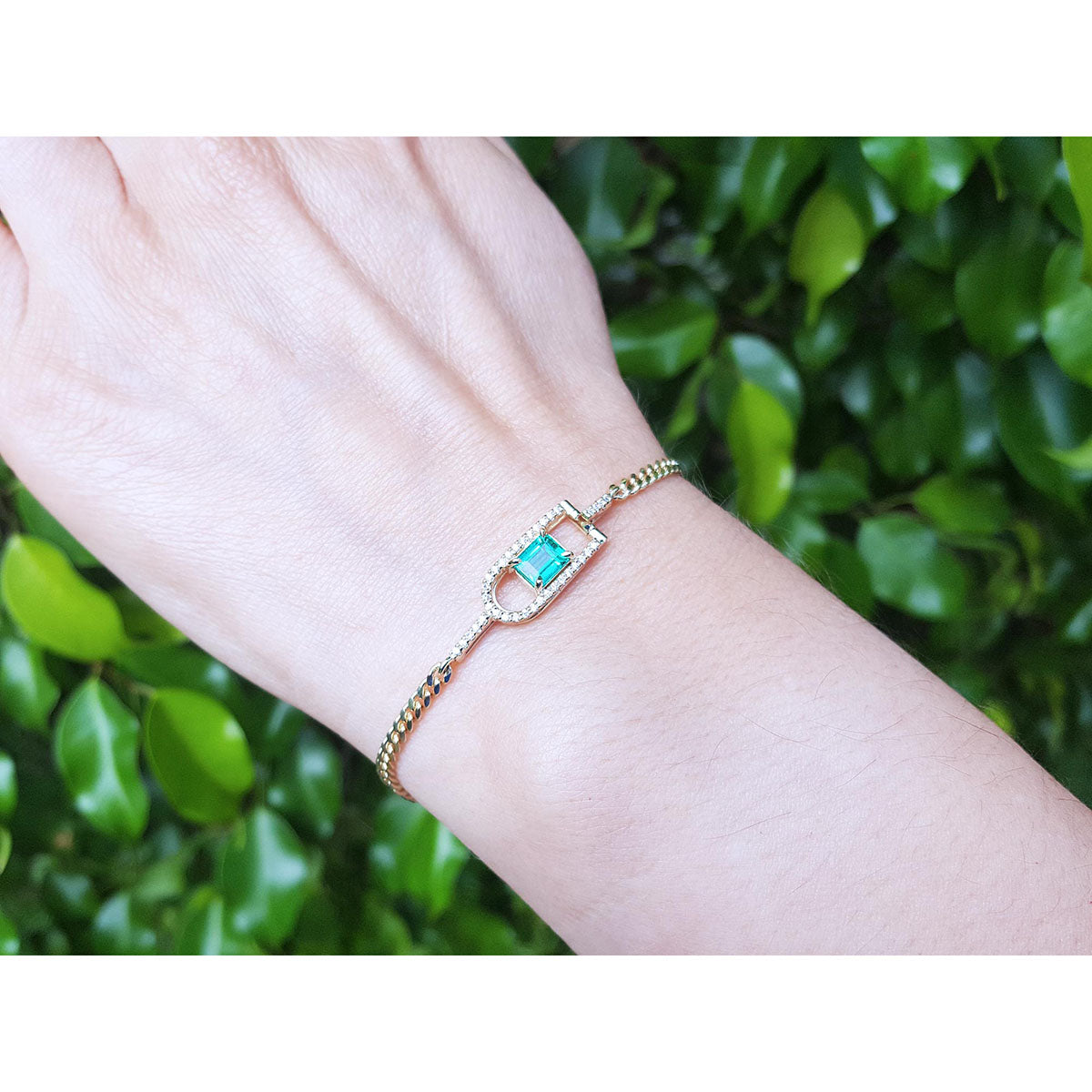 Gold bracelet with a square emerald on a wrist against a green leafy background.