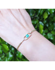 Gold bracelet with a square emerald on a wrist against a green leafy background.
