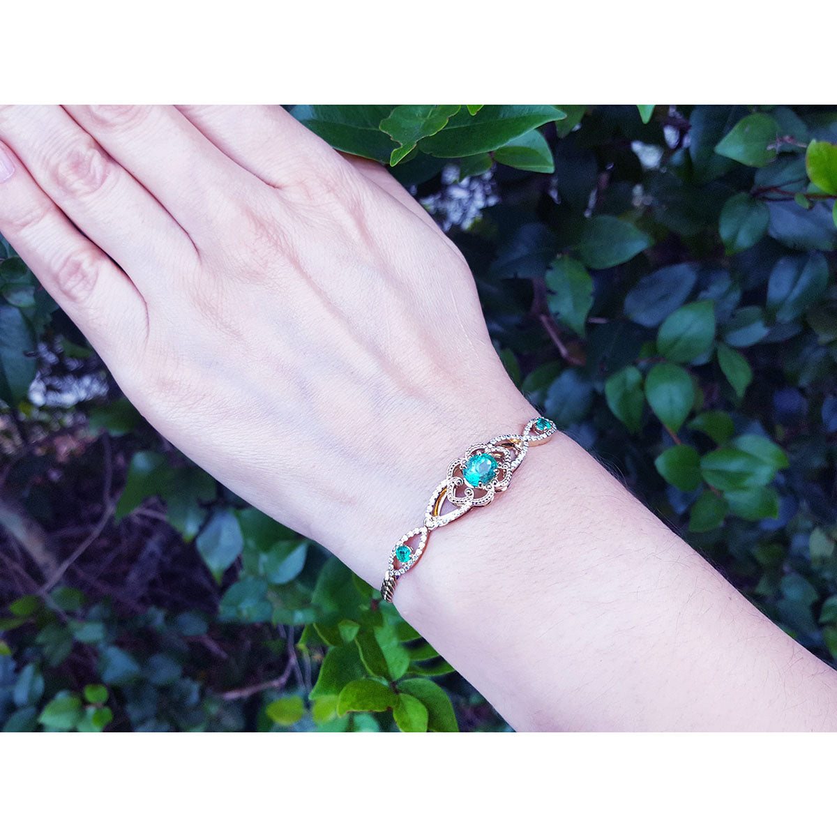 Hand wearing a bracelet with emeralds against a green leafy background.