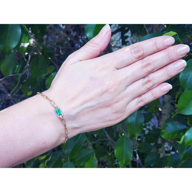 Woman's hand wearing a 14K yellow gold paper clip bracelet with a rectangular emerald and diamonds.