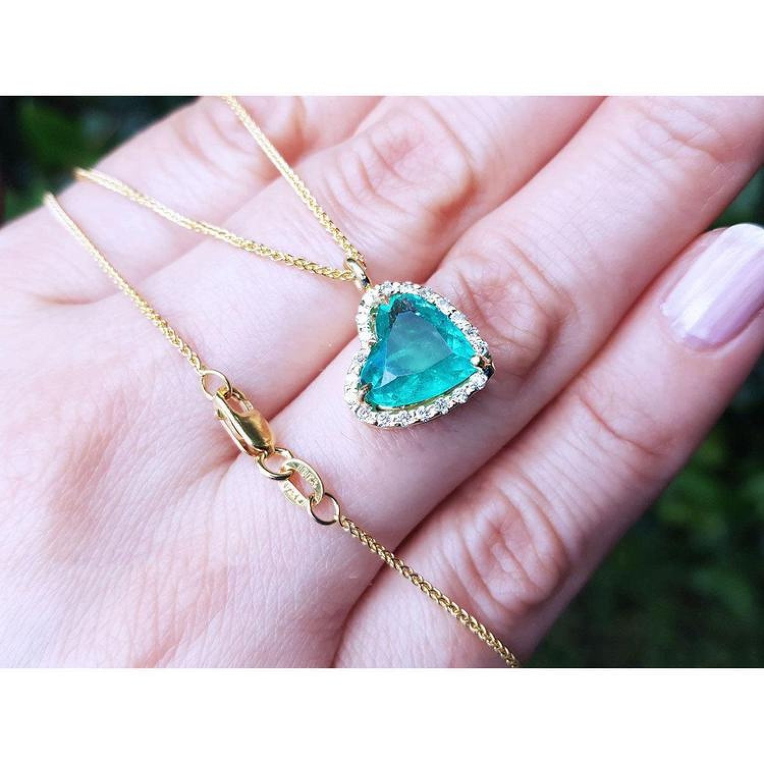 Tilted heart emerald necklace with halo diamonds in 18K yellow gold, shown on a hand.