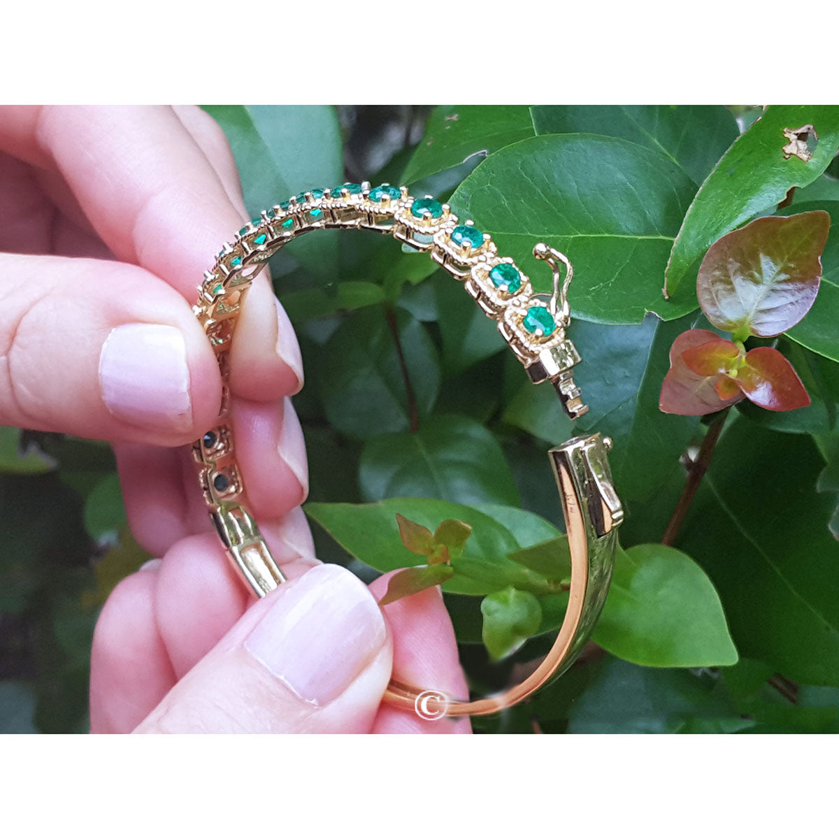 Gold bangle bracelet with round emeralds held by a hand between fingers against a leafy background