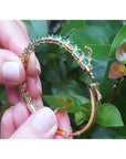 Gold bangle bracelet with round emeralds held by a hand between fingers against a leafy background