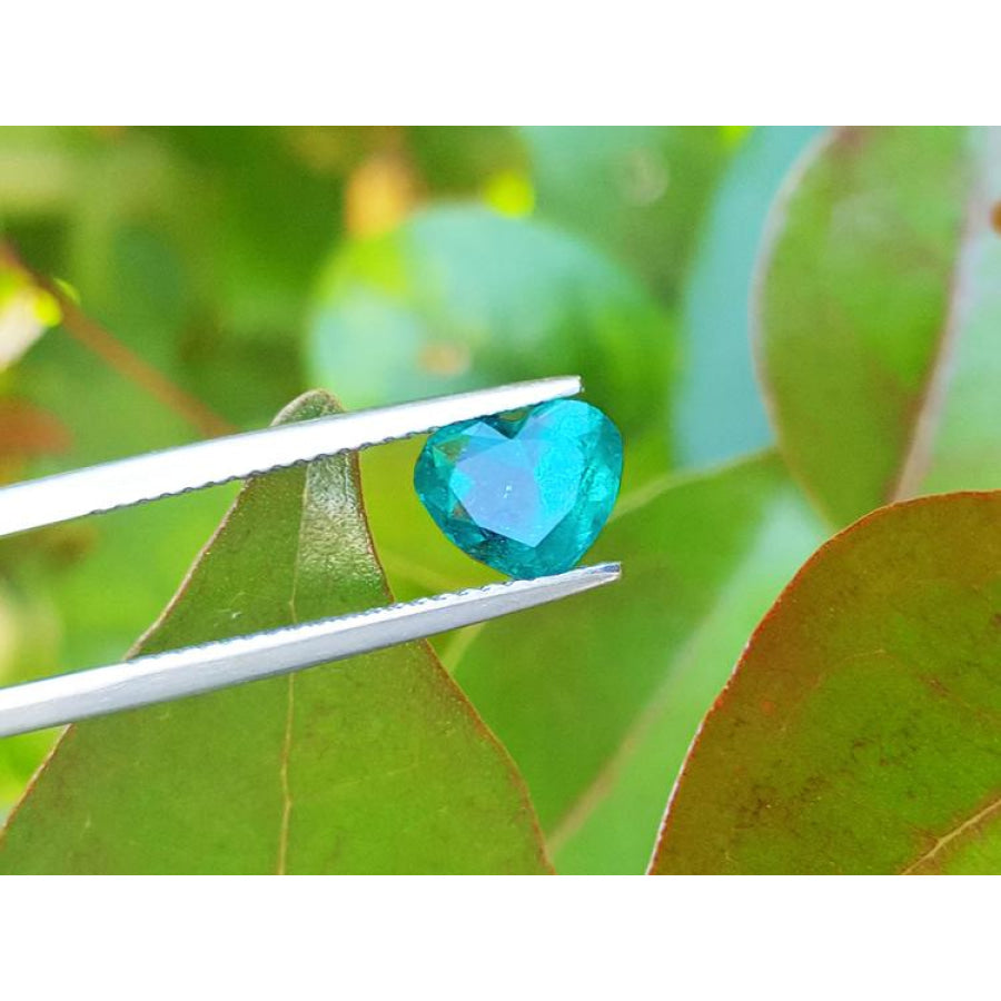 1.26 ct heart cut Colombian emerald, vibrant bluish-green, held by tweezers against green leaves.