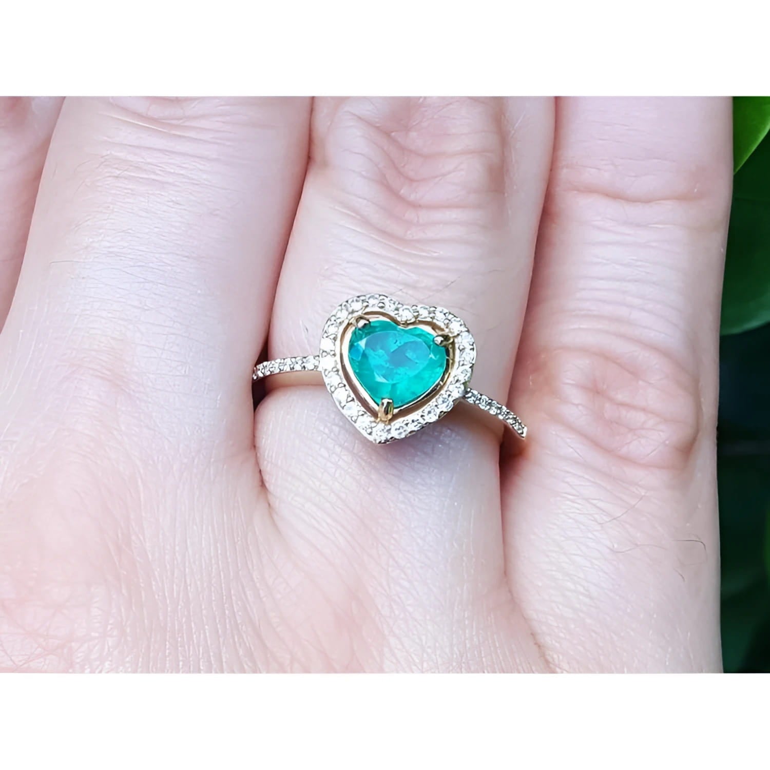 Heart-shaped emerald ring with halo diamond made of 14k yellow gold on a finger.