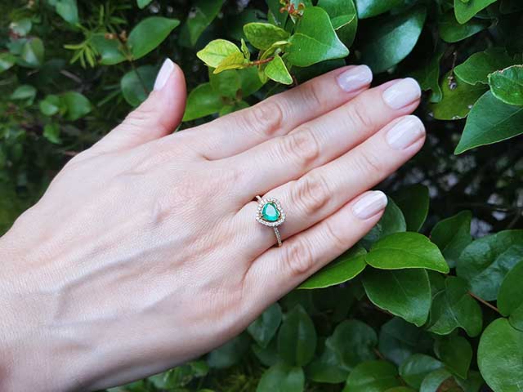 Hand wearing a heart cut emerald ring with a green leafy background.