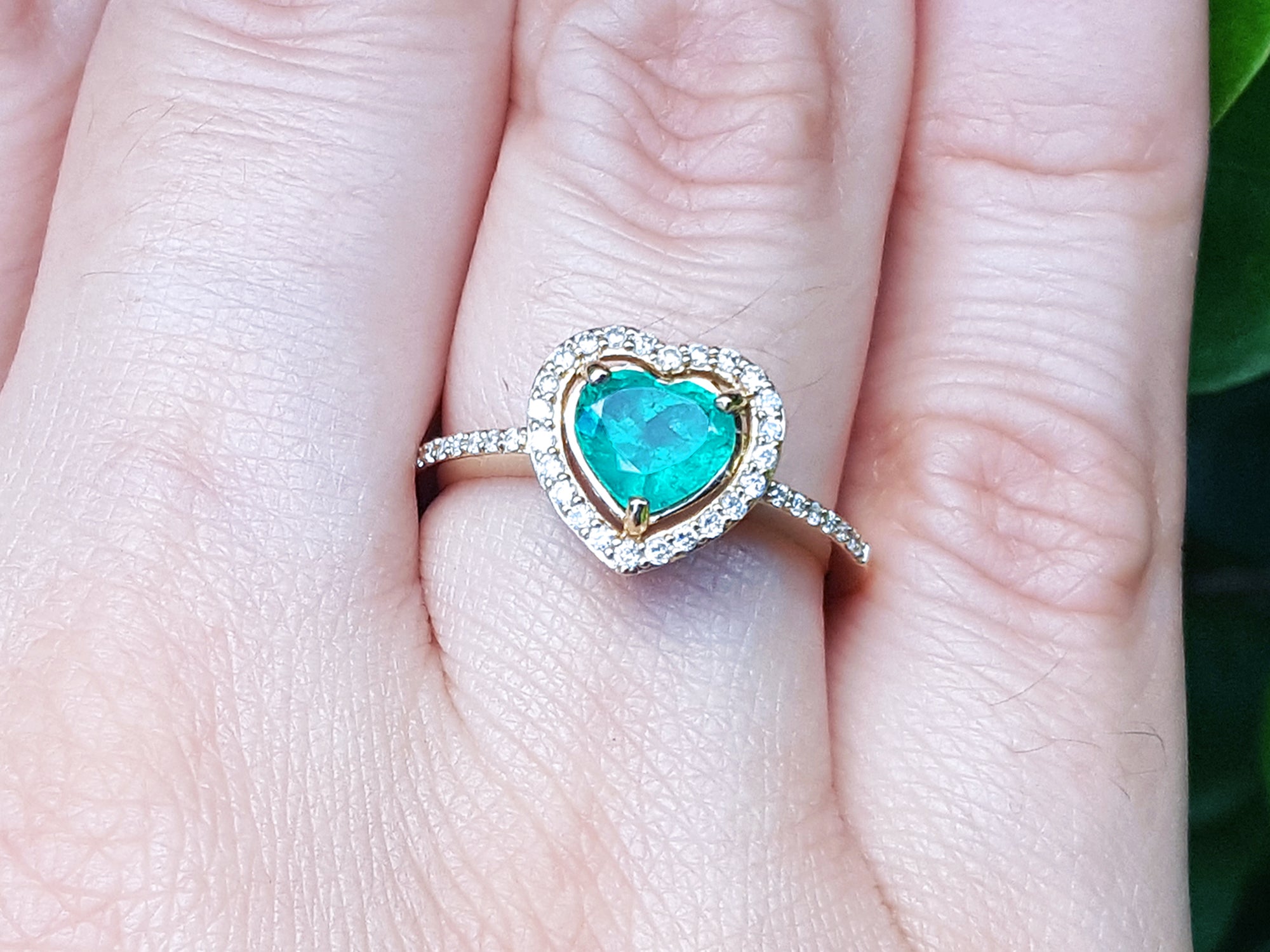 Heart-shaped emerald ring with halo diamond on a finger with a blurred green background.