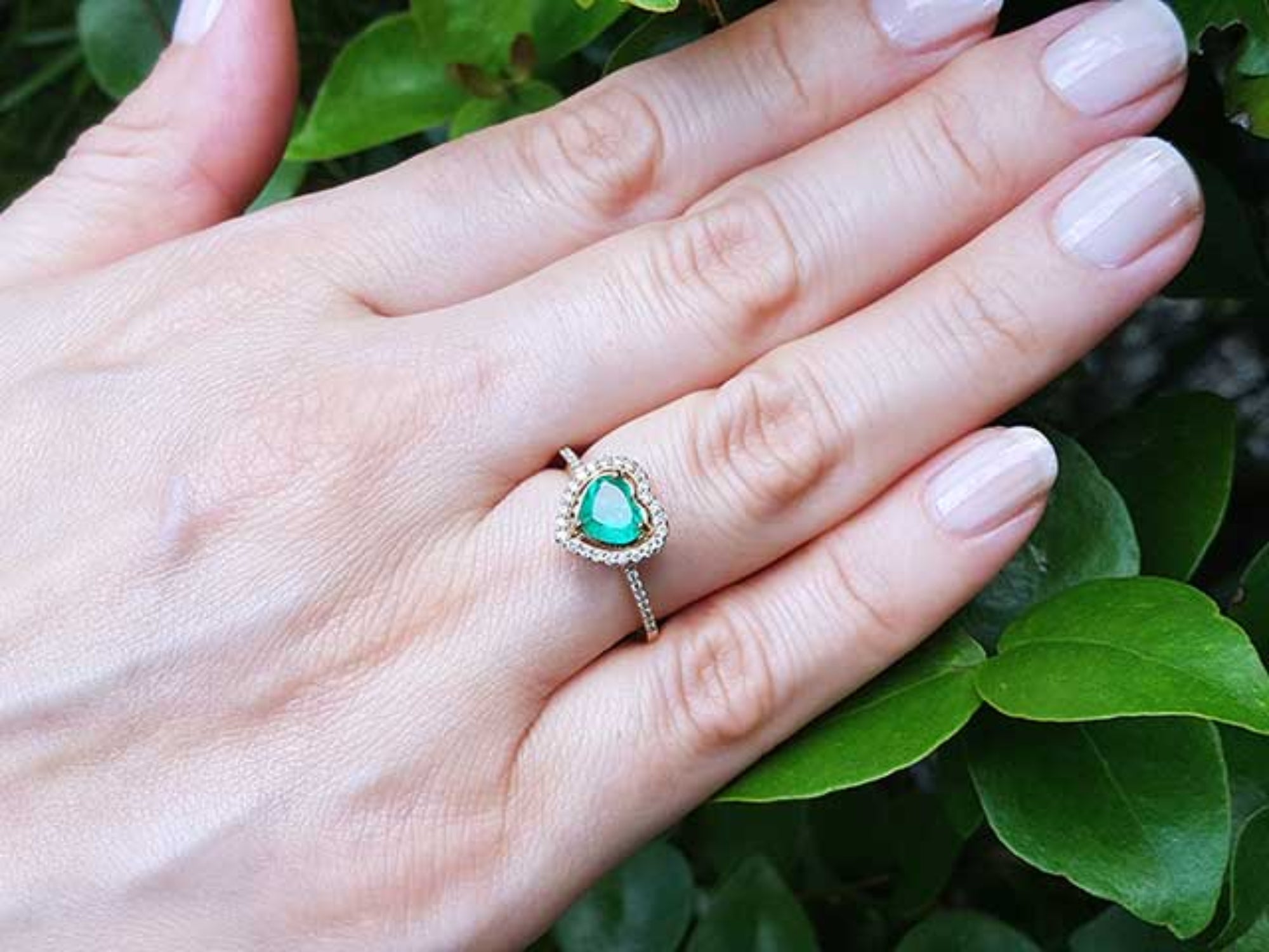 Hand wearing a ring with a heart cut emerald against a leafy background.