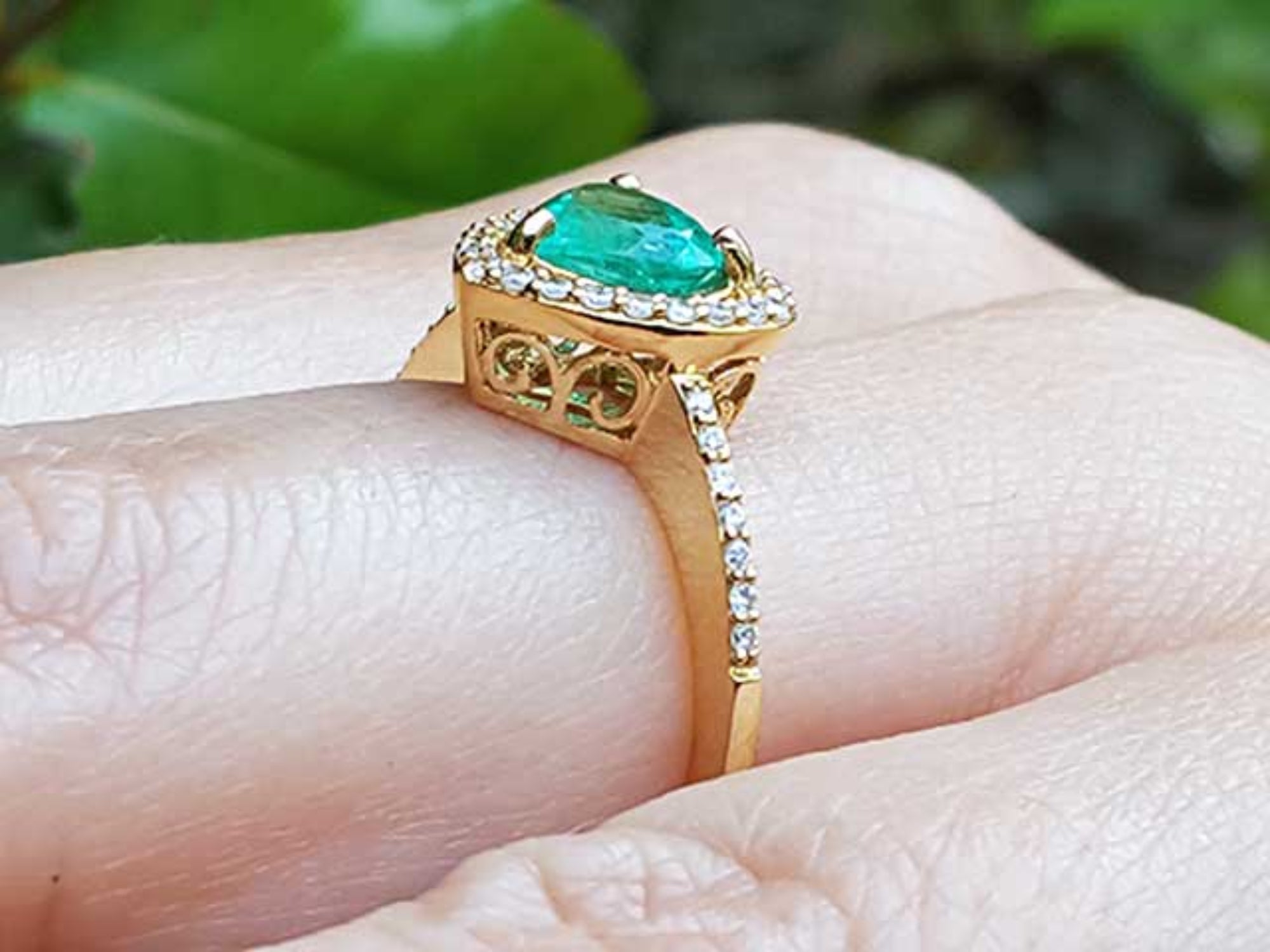 A 14k yellow gold ring with a natural emerald on a finger against a blurred natural background.