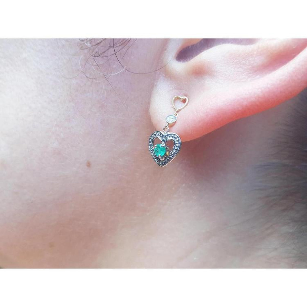 14K yellow gold heart earrings with brilliant cut Colombian emeralds, prong set, on an ear.