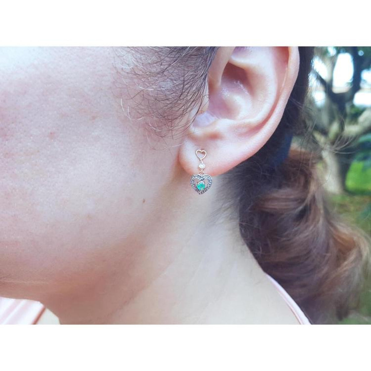 14K yellow gold heart earrings with brilliant cut Colombian emeralds, worn on an ear.