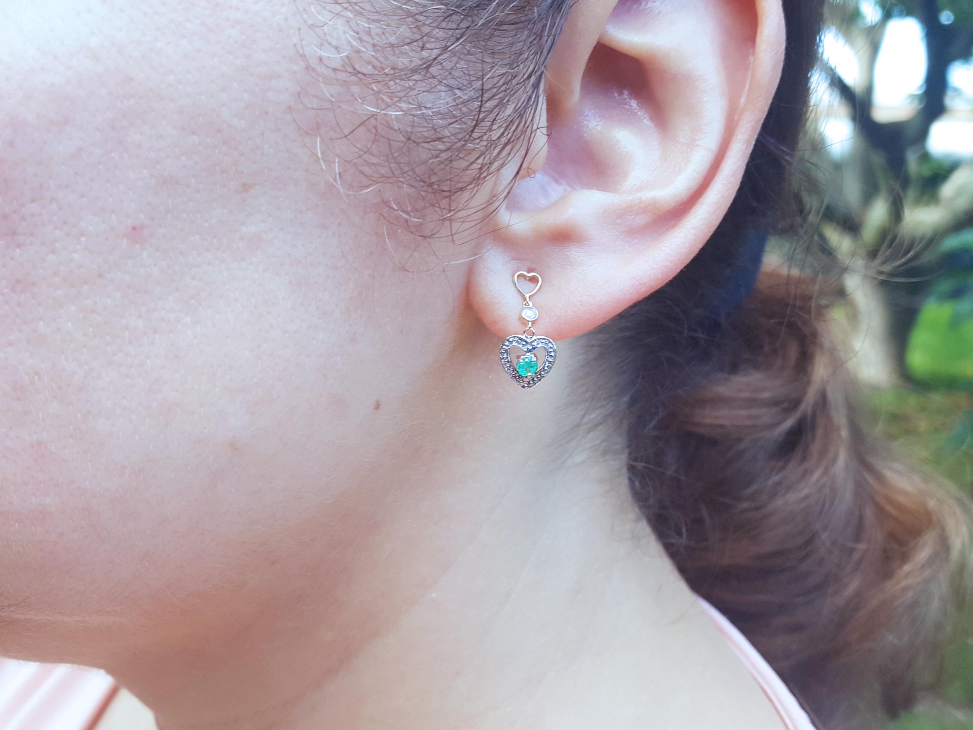 Woman&#39;s ear wearing a heart shaped earring with a round emerald.