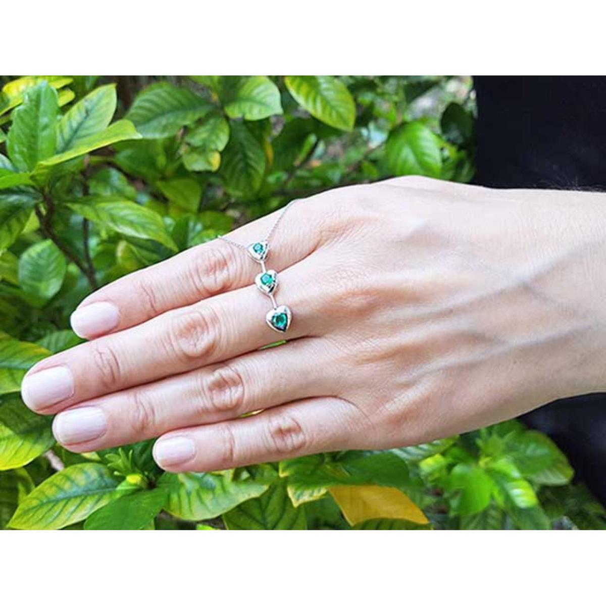 Hand displaying a three links heart shaped journey pendant with round cut emeralds against a green leafy background.