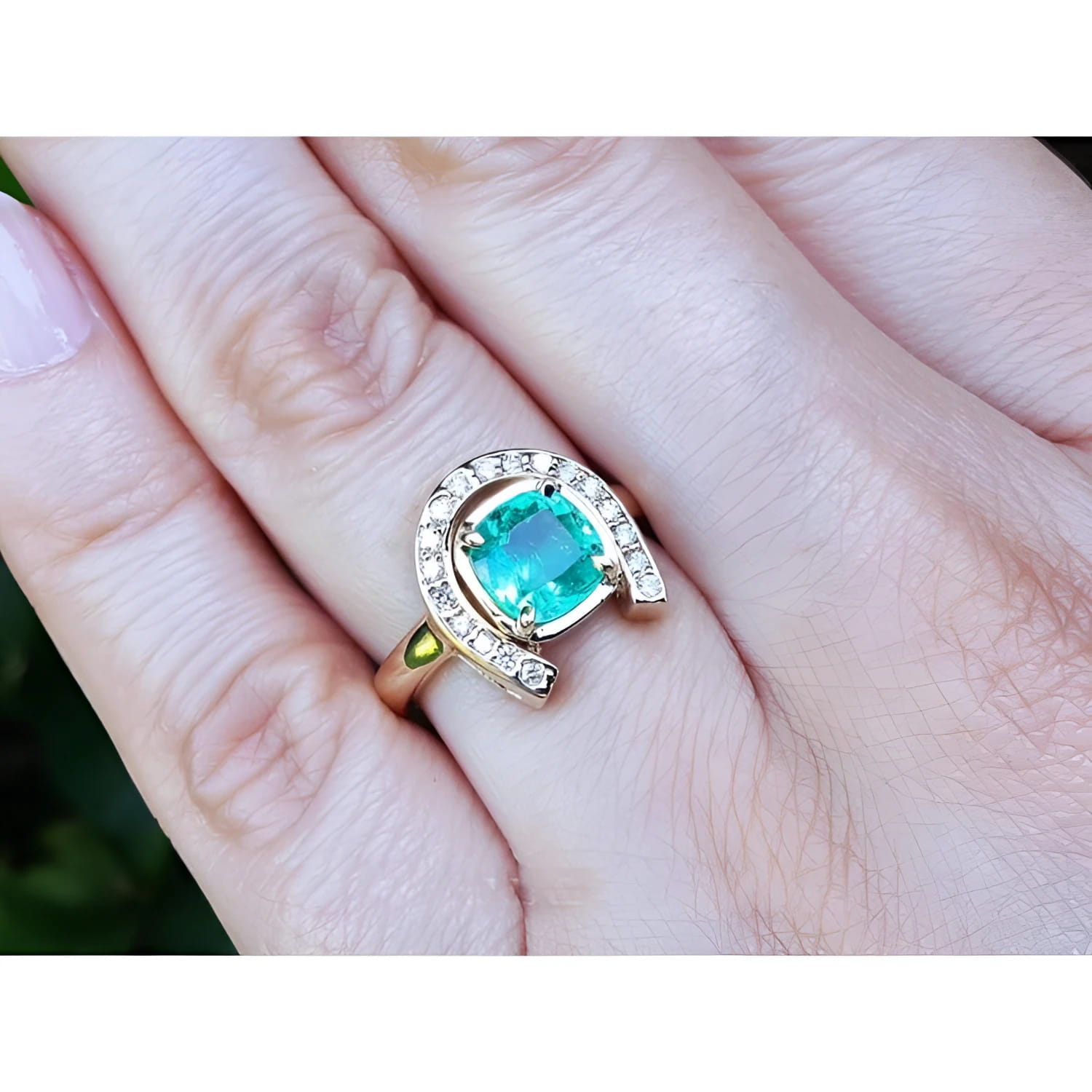 14K yellow gold horseshoe ring with a cushion-cut emerald and diamond halo, worn on a hand.