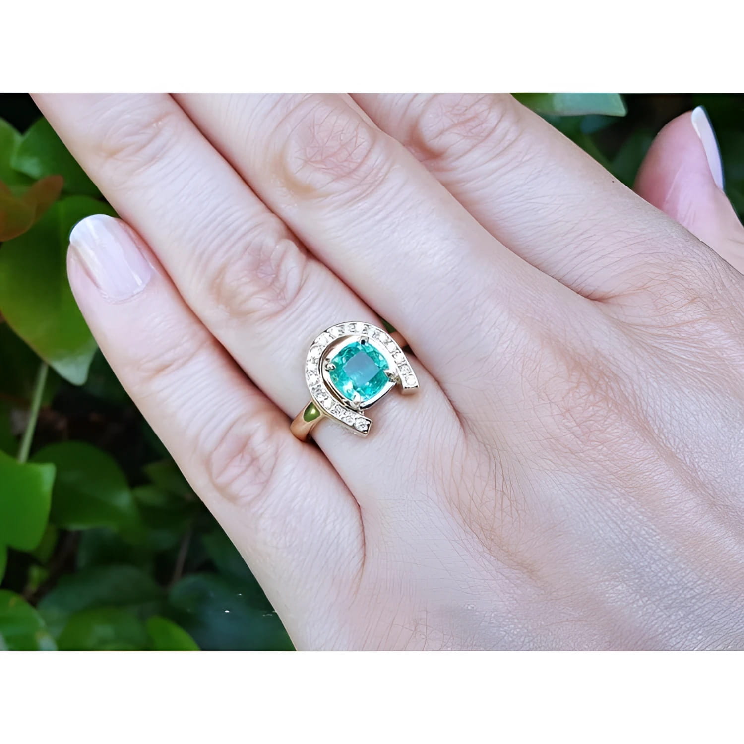 14K yellow gold horseshoe ring with cushion-cut emerald and diamond accents on a woman's hand.