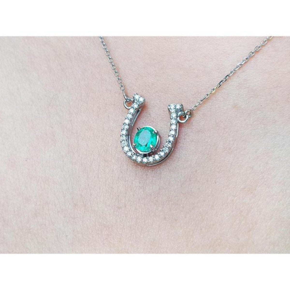 14K white gold horseshoe necklace with a prong-set oval emerald and natural diamonds.