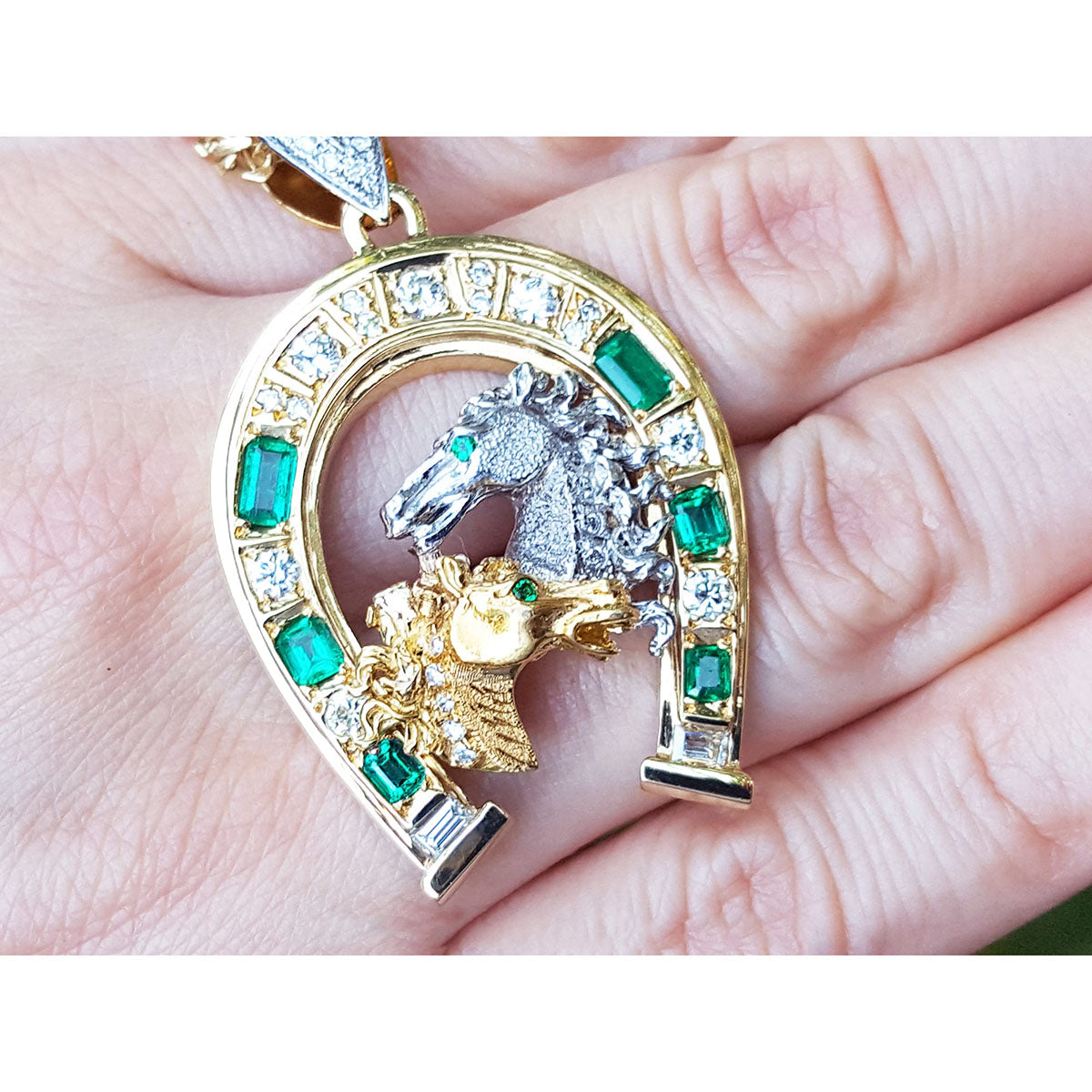 White and yellow gold pendant with horse design and emerald stones and diamonds held on a hand.