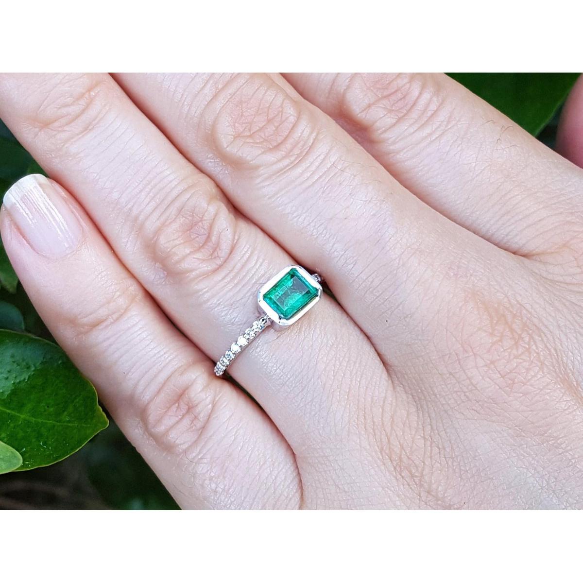 Hand wearing a bezel set emerald ring against a green leafy background.