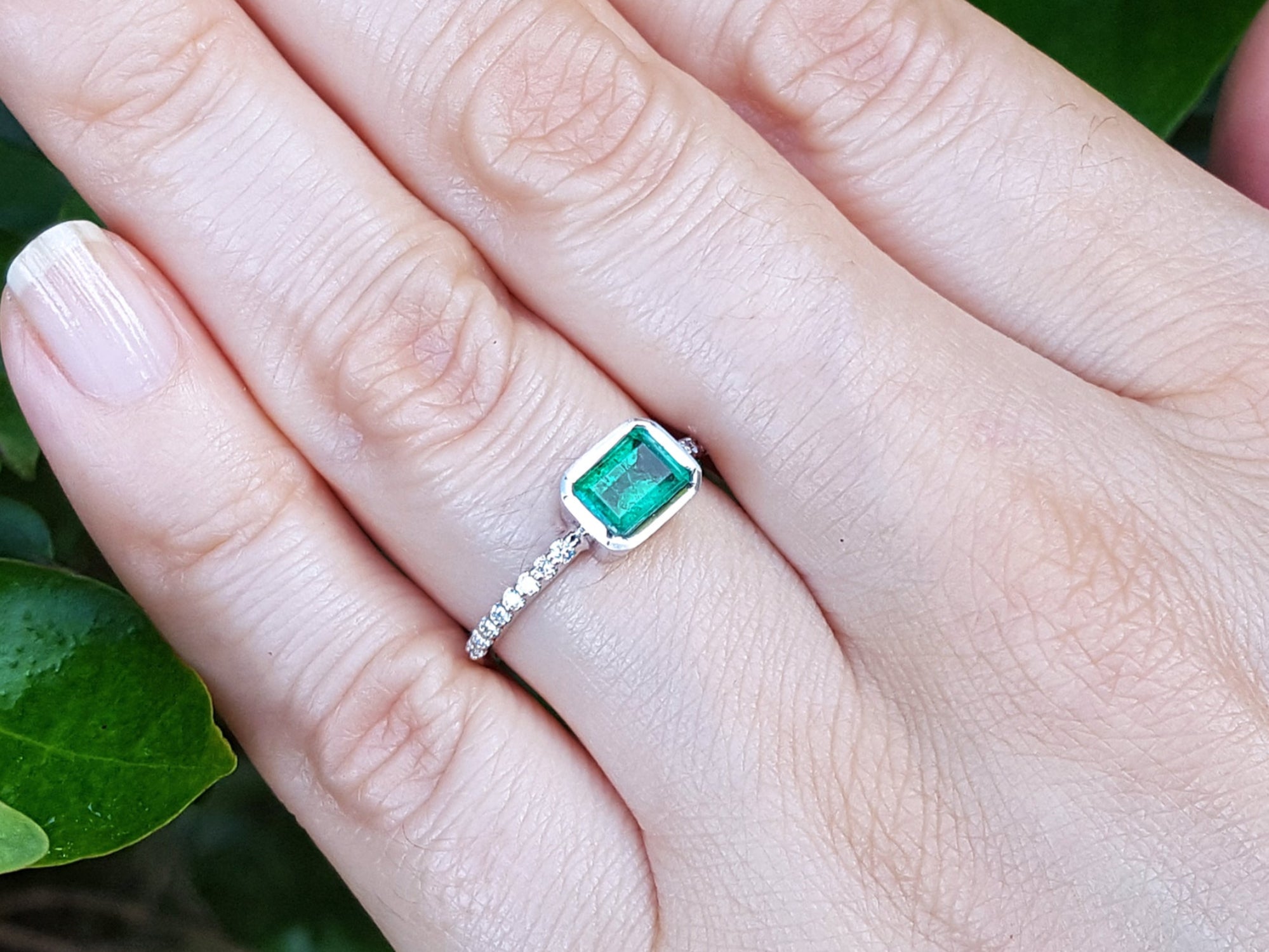 Hand wearing a bezel set emerald ring against a green leafy background.