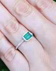 Hand wearing a bezel set emerald ring against a green leafy background.