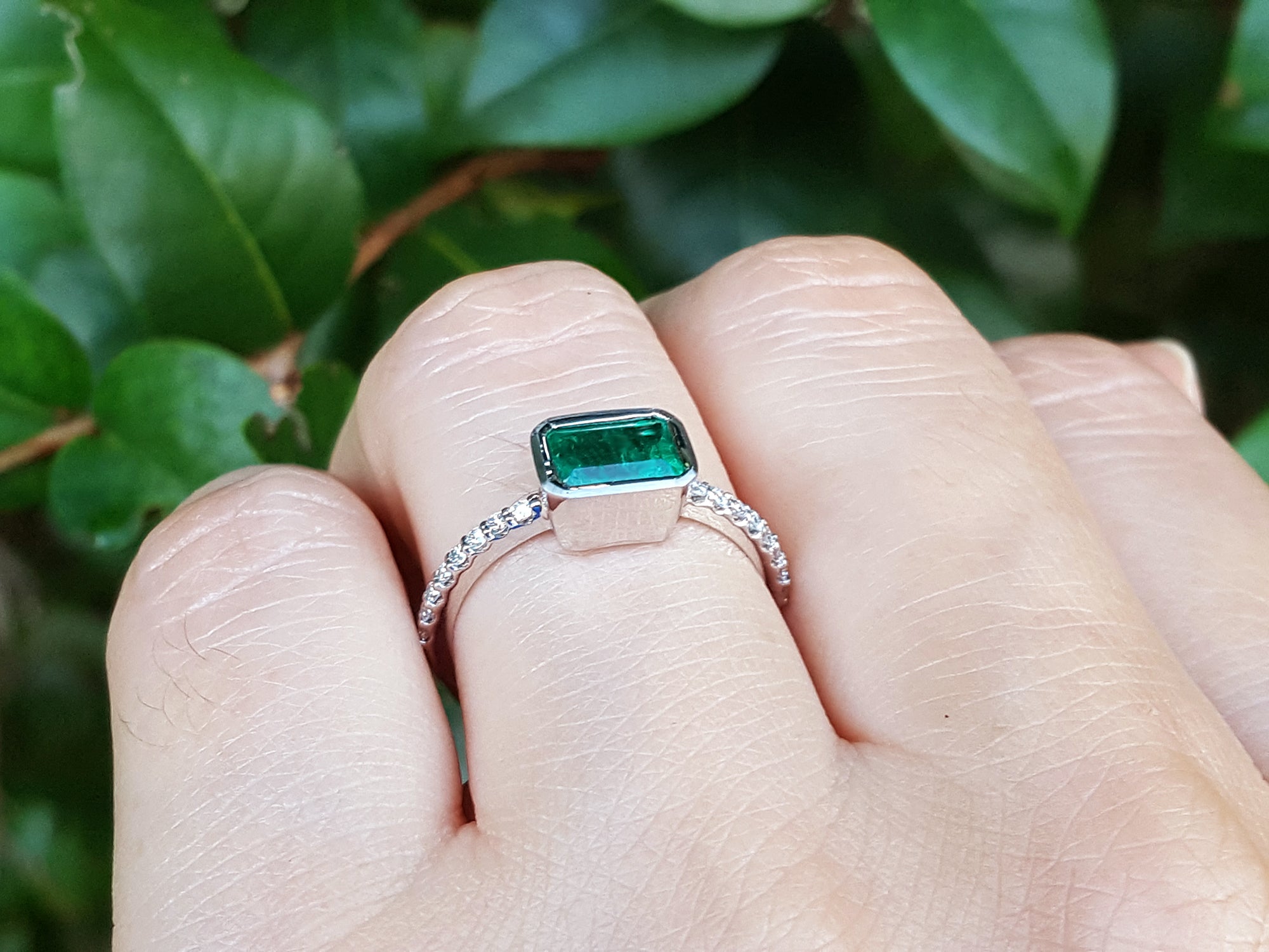 A white gold emerald ring on a finger against a blurred green leafy background.