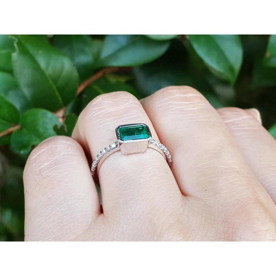 0.73 ct bezel set emerald ring with accent diamonds on a 14K white gold band, worn on a finger.