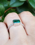 A white gold emerald ring on a finger against a blurred green leafy background.