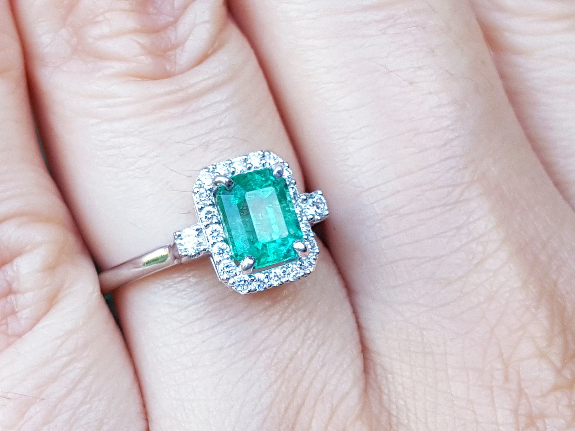 An inexpensive emerald engagement ring with a halo diamond on a finger.