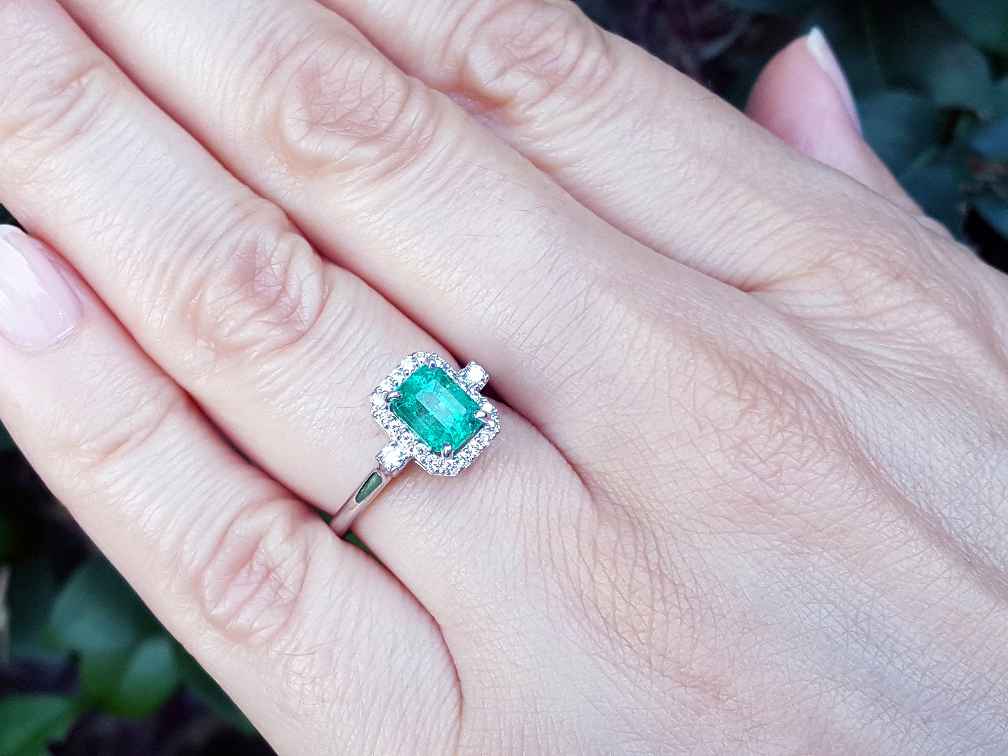 An emerald engagement ring with halo diamond on a hand with a natural background.