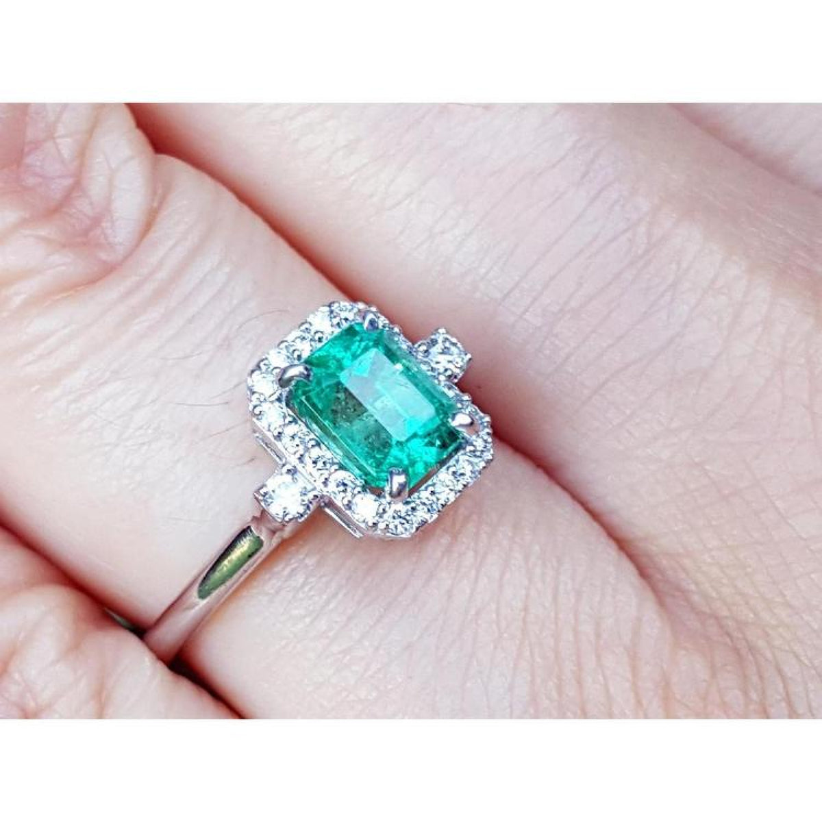 1.47 ct emerald cut emerald ring with diamond halo in 14K white gold on a finger.