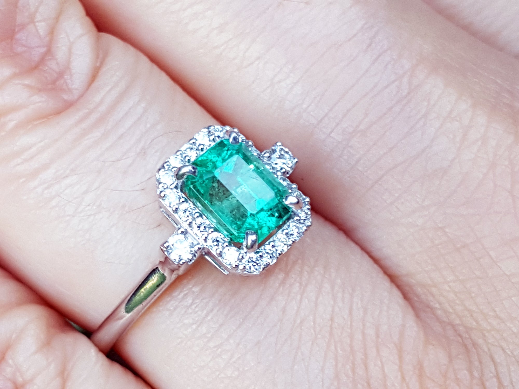 This inexpensive emerald engagement ring with halo diamond is on a finger.