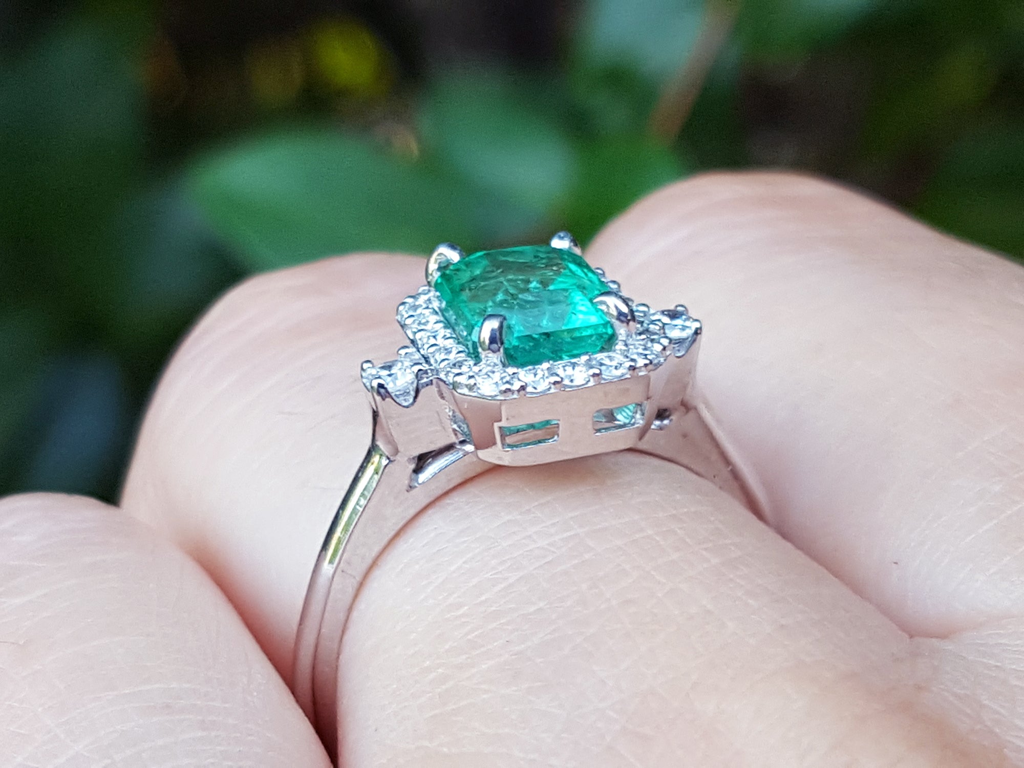 The underside view of an emerald engagement ring between fingers against a blurred natural background.
