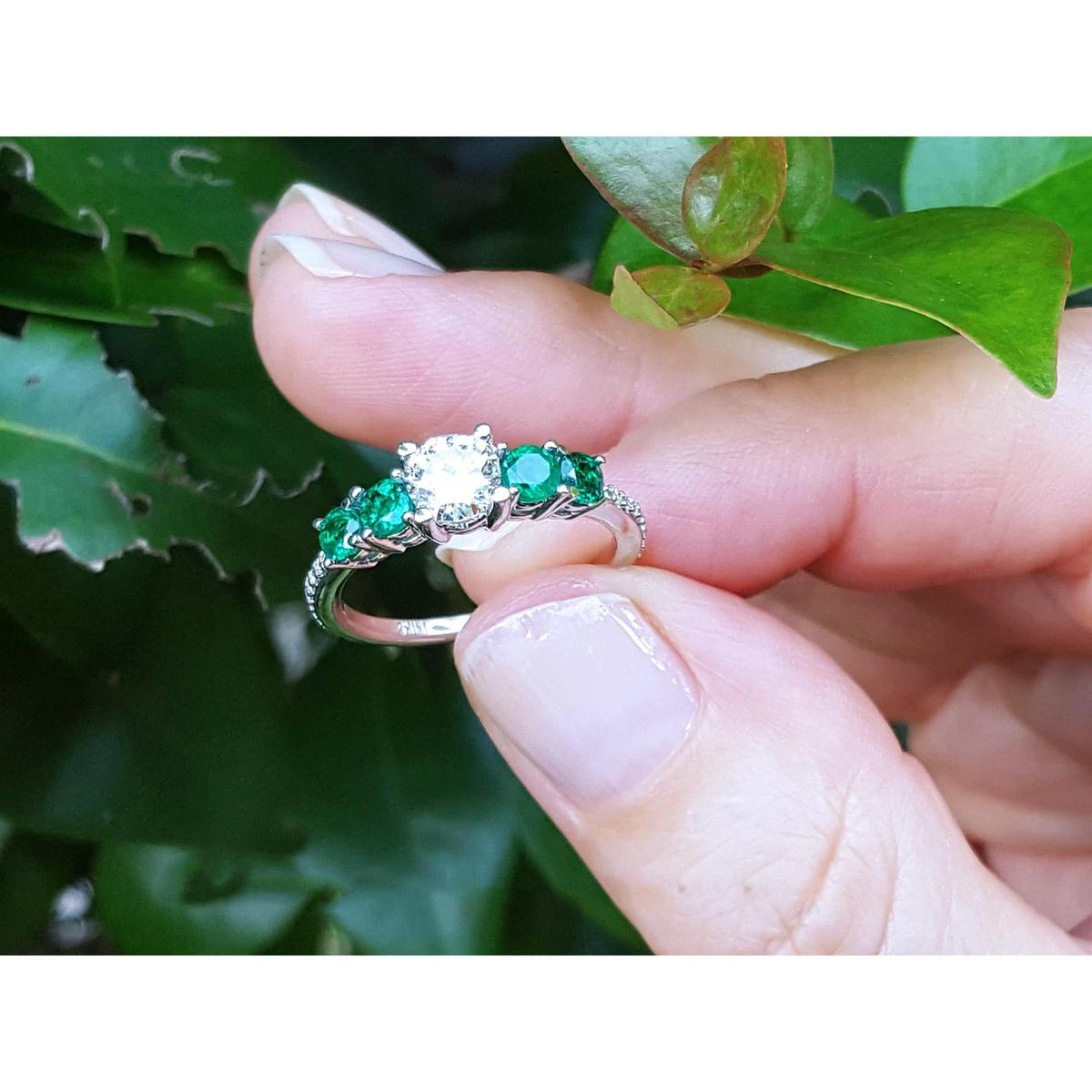 A white gold ring with emeralds held between fingers against a green leafy background.